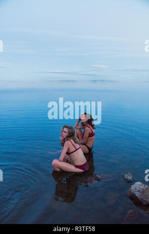 Two young women wearing bikinis, sitting on a stone in a lake Stock Photo