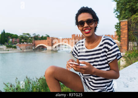 Smiling young woman wearing sunglasses at the riverside using cell phone Stock Photo