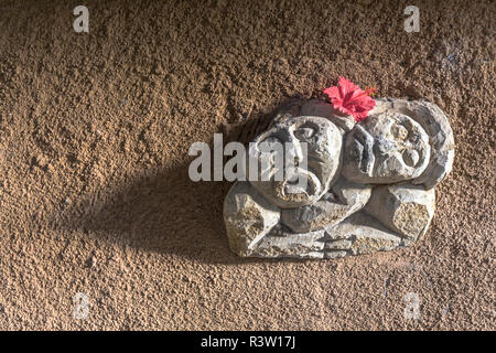 Two sculptured faces on a very rough wall in Bali with a red flower on top and dramatic lighting. Stock Photo