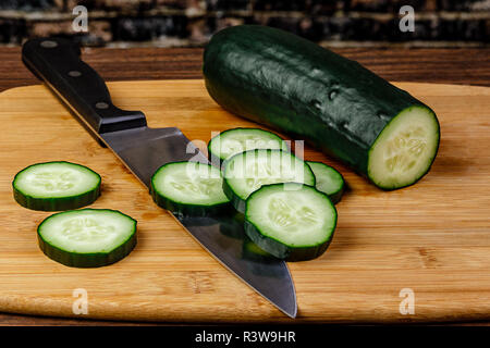 Slicing Cucumbers with a sharp Chef's knife on a bamboo cutting board with a vintage brick wall in the background. Stock Photo