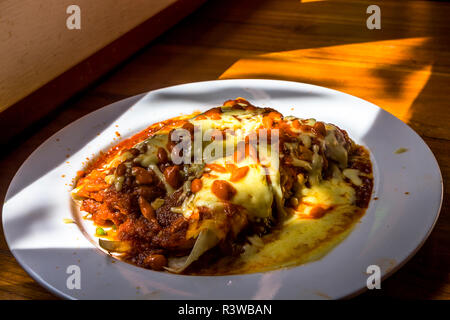 Enchilada’s served with cheese, chili sauce filling a white plate with high lighted areas and shadows on a wood table. Stock Photo