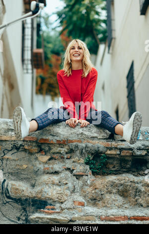 Happy young blond woman sitting on urban background. Smiling blonde girl with red shirt enjoying life outdoors. Stock Photo