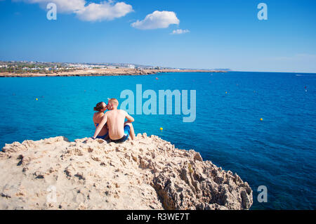 Romantic couple sitting together on a rock near transparent water, kissing against several clouds in the sky. Concept of love, care, tenderness, trave Stock Photo