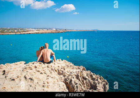 Romantic couple sitting together on a rock near transparent water. Woman leaning her head on man's shoulder. Concept of love, care, tenderness, travel