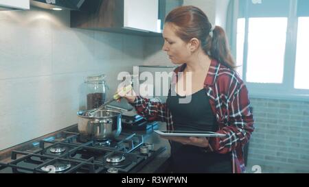 woman cooks soup in the kitchen prevents the water in the pot and looks at the recipe on the digital tablet. woman in the kitchen slow motion video. girl mom in the kitchen indoors preparing food concept smart home control on a digital tablet lifestyle Stock Photo