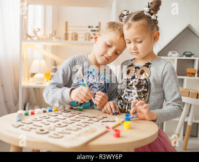 Preschooler girl and boy play and draw by colorful paints next to English wooden alphabet. Stock Photo