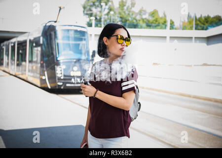 Pretty brunette woman smoking an e-cigarette standing in a street exhaling a cloud a smoke from her mouth while looking off to the side Stock Photo