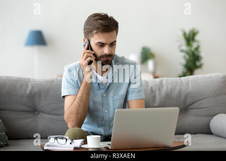 Serious man talk on smartphone using laptop at home Stock Photo