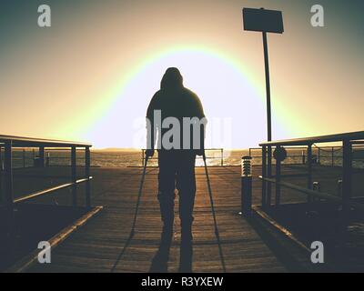 Bended disabled man on coast mole rest on his forearm crutches, warm sunset in background Stock Photo