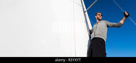 Yacht captain with a beard stands on sail boom on a sailing yacht, holding the rope in his hand and smiling, feeling happy. Adult yachtsman travelling around the world. Copy space Stock Photo