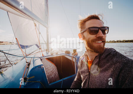 Man dressed in casual wear and sunglasses on a yacht. Happy adult bearded yachtsman close-up portrait. Handsome sailor on a boat smiling during regata on a sea or river. Stock Photo