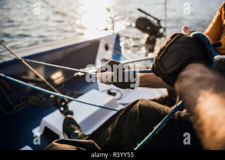 Close-up of male hands in gloves pulling rope of sailboat while mooring yacht and sitting with friends on deck. Stock Photo