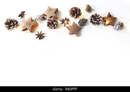 Christmas festive border, banner. Winter arrangement of pine cones, anise, wooden stars and glowing Christmas lights chain on white table background. Flat lay, top view. Empty space. Stock Photo