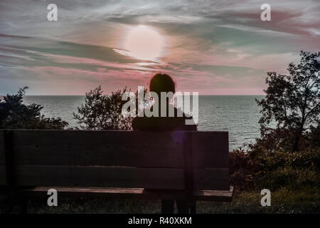 A woman, seen from behind, sitting on a bench, gazing  the sunset on Lake Huron. Stock Photo