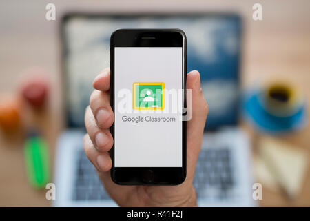 A man looks at his iPhone which displays the Google Classroom logo, while sat at his computer desk (Editorial use only). Stock Photo