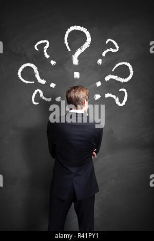 businessman having many questions symbolized as questionmarks on a blackboard over his head Stock Photo