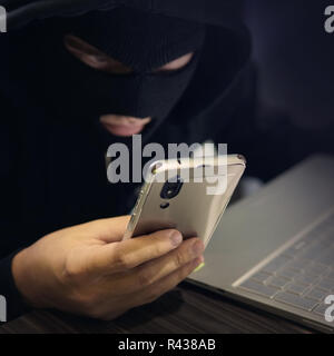 Male hacker in a black mask uses smartphone and laptop. A fraudster commits cyber crime. Fraudulent scheme with personal data and banking. Attempt to  Stock Photo