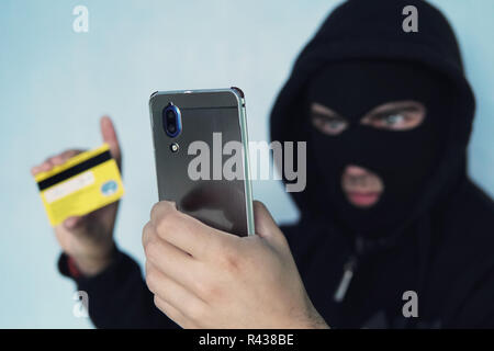 The thief scammer is holding a mobile phone and credit card. The concept of Bank card fraud through a virus in the smartphone. a thug in a black Balac Stock Photo