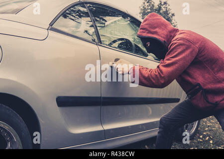 Side view of car being forced by a man in hoodie and mask. Thief tries to steal vehicle from a parking. Young male acts alone breaking the car door. U Stock Photo