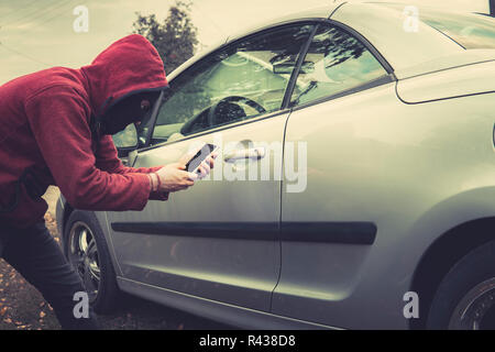 Side view of young criminal in black balaclava and hoodie holding smartphone and trying to interact with car by mobile application. Unknown person tri Stock Photo