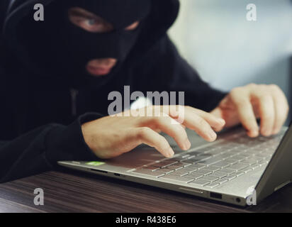 Close up of a ill-intentioned hacker in black balaclava entering the data on a laptop. Computer hacker commits cyber crime. A sitting man with hidden  Stock Photo