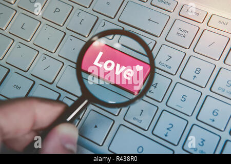 Written word love on keyboard red button viewed through the magnifying glass. Close up of male hand with loupe searching for love in internet. Concept of love search online. Dating services in net. Stock Photo