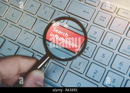 Button Search information on the laptop keyboard viewed through the magnifying glass. Man holds the loupe above the computer keypad with the red key search. Concept of using online search engines. Stock Photo