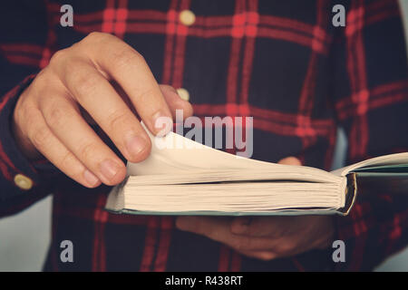 Close up of a young male in a chequered shirt holding the book and turning over the page. Ingusitive man reads the book on the run. Person is standing and reading. Cursory reading. Leafing over pages. Stock Photo