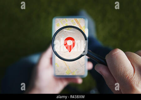 Shot of male hands holding magnifying glass with the red icon of geo-location and looking through it to the smartphone with online city map on the screen. Concept of using mobile net for navigation. Stock Photo