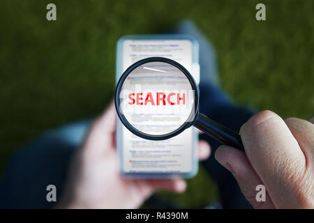 Close up of a man sitting and looking through magnifying glass with red written word Search on the lens to the smartphone screen. Using mobile internet in searching information. Browsing in cellphone. Stock Photo