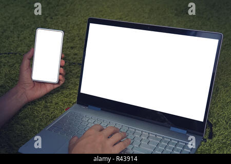 Close up of man lying on soft green surface and using digital devices - modern laptop and smartphone with white blank screens. Man types on the laptop keyboard and looks at the cellphone. Copy space. Stock Photo