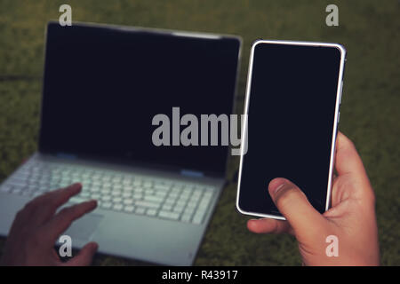 Image of man's hand holding cell phone with black screen and blur silver laptop on green soft surface as background. Usage of gadgets at home. Man interrupts laptop work for answering phone call. Stock Photo