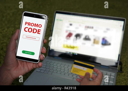 Close up of male hands holding smartphone with red written words Promo code on the screen and credit card on blur laptop background. Online shopping website on laptop. Easy e-commerce using gadgets. Stock Photo