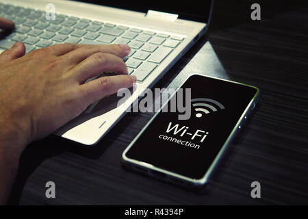 Close up of male hands typing on laptop keypad and checking the smartphone wi-fi connection. Cellphone with signal icon and written words WI-FI connection on black screen near the personal computer. Stock Photo