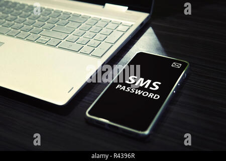 Table with open laptop and black-screened smartphone with white written words SMS password on its display. Concept of linking the work of computer and mobile via sms password entering. One time code. Stock Photo