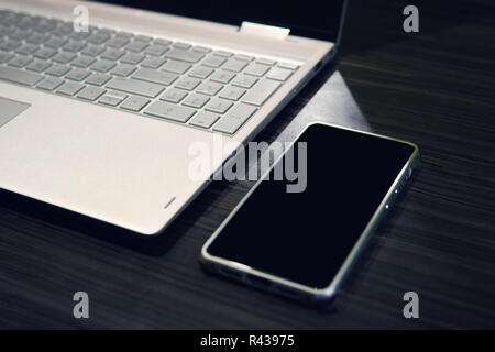 Smart phone with copy space screen and laptop on wooden table background. Empty black screen of a mobile left on the table near the computer. Use of digital gadgets at work. Phone display mockup. Stock Photo