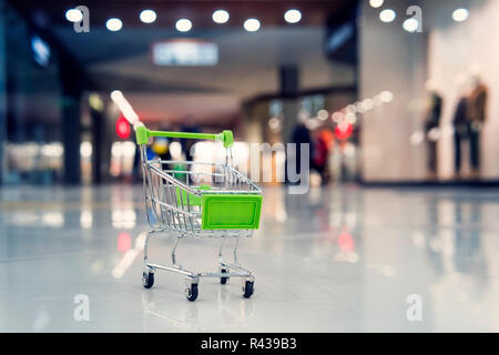 A small green toy cart in a large shopping Mall. The concept of shopping and sales in supermarkets.. Blur shopping cart in superstore. Stock Photo