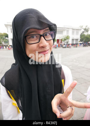 Vertical portrait of Indonesian teenager wearing black headscarf and white dress and glasses with grin making peace sign in Jakarta square. Stock Photo