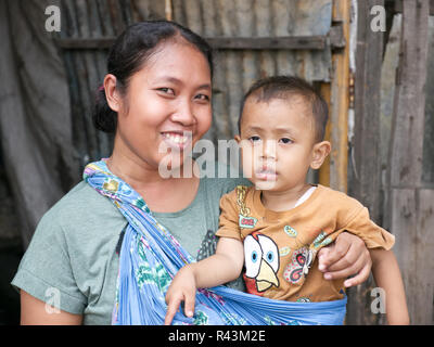 Horizontal portrait of smiling, proud, happy young Indonesian Mother holding young boy wearing yellow t-shirt in front of wood and tin home. Stock Photo