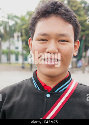 Vertical portrait of young Indonesian teenage boy wearing black jacket with red and white strap across chest looking at camera and smiling. Stock Photo