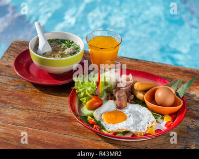 Breakfast set, fried egg, soft-boiled rice, orange juice on wooden table with blue background of water in pool. Stock Photo