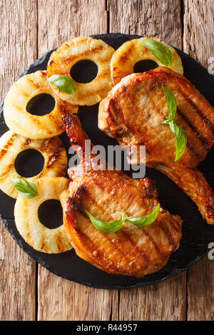 Grilled pork chop in honey glaze served with pineapple and basil close-up on slate on the table. Vertical top view from above Stock Photo