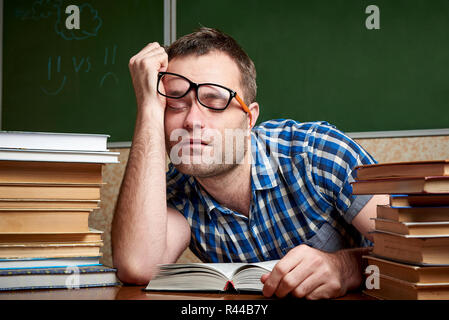 A tired and tortured disheveled student in glasses is sleeping at a table with stacks of books against the background of a cool blackboard. Stock Photo