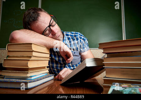 Tired and tortured disheveled student in glasses is sleeping at the table on a stack of books against the background of the chalkboard. Stock Photo