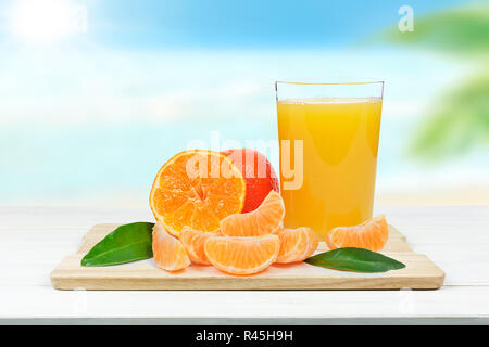 Glass of fresh orange juice and orange or tangerine fruits with leaves on blurred background of a tropical beach (mixed) Stock Photo