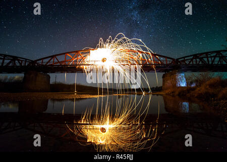 Light painting art concept. Spinning steel wool in abstract circle, firework showers of bright yellow glowing sparkles on long bridge reflected in riv Stock Photo