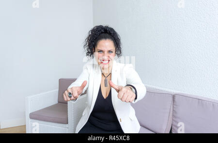 Portrait of a middle age 40s woman with blue eyes, white skin and dark hair looking at the camera and smiling with red lipstick making thumbs up gesture with both hands. Wearing white blazer Stock Photo