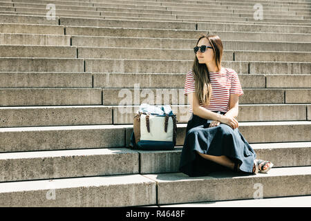 Beautiful tourist girl or student in sunglasses and with a backpack sits on the stairs and rests. Lifestyle Stock Photo