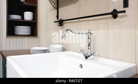 Light kitchen in classic style with a luxury mixer and ceramic sink, with dishes on the shelves in a wooden cupboard, rustic style Stock Photo