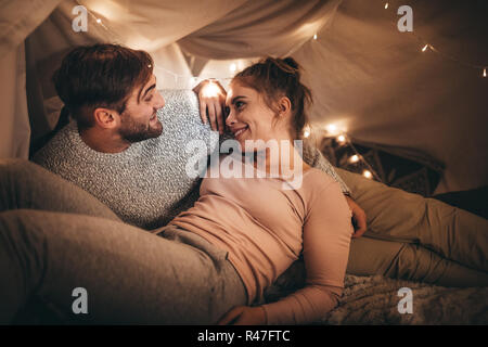 Happy man and woman lying on bed talking while looking at each other. Romantic couple spending time together talking on bed at night with tiny light b Stock Photo